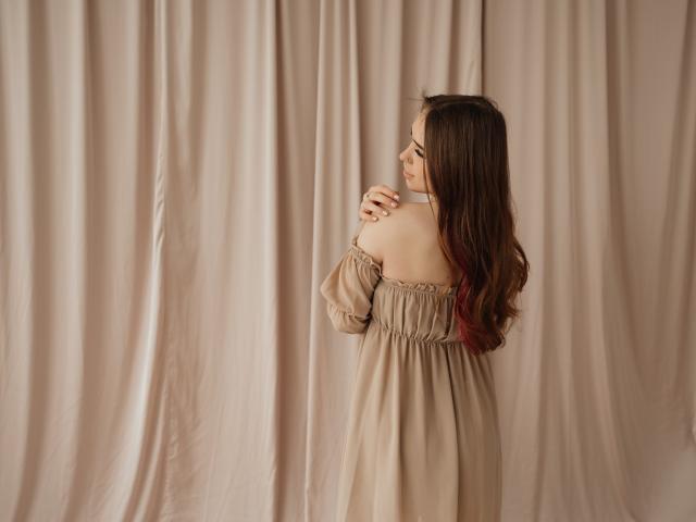 LauraHlot, con el pelo largo y castaño oscuro, está de espaldas, vestida con un vestido beige de corte asimétrico, con las manos juntas cerca de su cuerpo. mentón. Está frente a un fondo de cortinas suaves y beige. Increíblemente sexy..