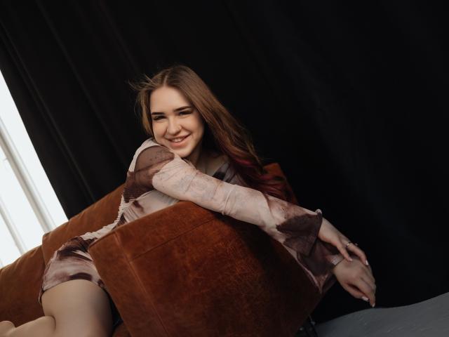 LauraHlot, avec de longs cheveux bruns, souriante, appuyée sur un canapé en velours marron, portant une robe marron et blanche à motifs. fond sombre, lumière du soleil venant de la gauche, pose détendue, légère tension sexuelle..