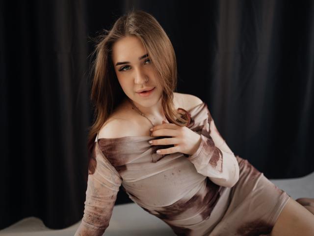 LauraHlot, een vrouw met bruin haar en lichte huid, draagt een strak, bruin jurkje met open schouders, leunt voorover en heeft haar hand op borst, seksuele spanning, verleidelijke blik, donkere achtergrond, zachte belichting, nonchalante pose.