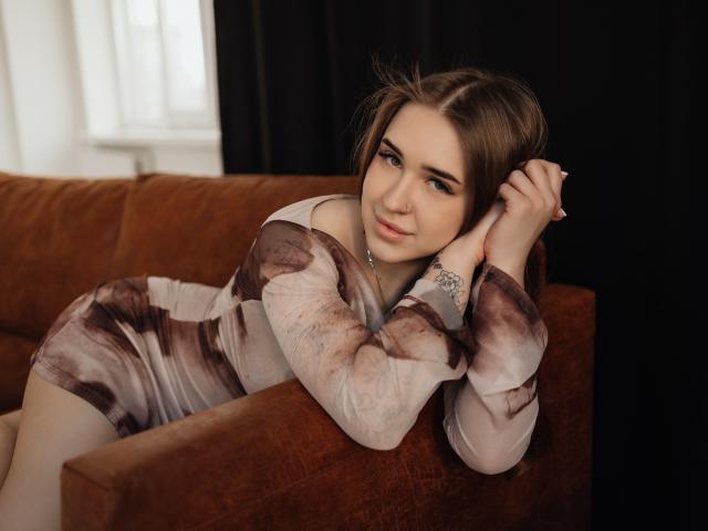 LauraHlot lehnt sich auf ein braunes Sofa, trägt ein durchsichtiges, braunes und beiges Kleid und blickt direkt in die Kamera. verführerisches Lächeln. Ihr dunkles Haar ist hochgesteckt, wodurch ihre durchdringenden Augen zur Geltung kommen..