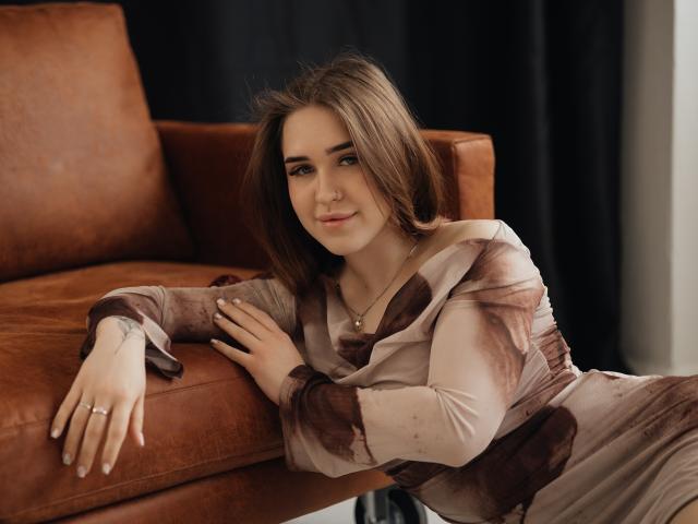 LauraHlot lehnt verführerisch auf einem braunen Ledersofa, trägt ein durchsichtiges, braun gefärbtes Kleid und legt ihre Hand auf den sitzt, langes, braunes Haar, sanftes Lächeln, Schmuck an den Fingern, sexy Ausstrahlung.