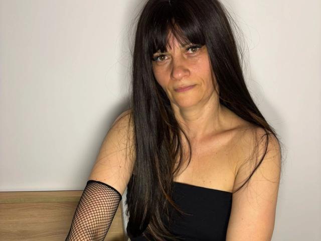 Photograph of LaraaFox-hot, a middle-aged woman with long black hair, bangs, and a black strapless top, wearing a fishnet arm sleeve. She's sitting against a white wall, looking seductively at the camera.