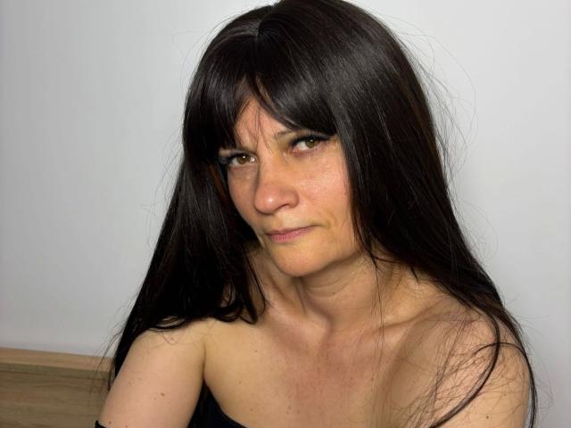 Fotografie von LaraaFox: Eine attraktive, mittlere Frau mit langen, schwarzen Haaren, braunen Augen und heller Haut, die direkt in die Kamera schaut. Kamera, mit einem leichten Grinsen, oben ohne, wobei die Schlüsselbeine sichtbar sind, leichte sexuelle Spannung..