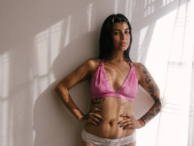 Fotografia di PsychesLoli in piedi contro un muro bianco, con la luce del sole che proietta ombre. Indossa un reggiseno di pizzo rosa e biancheria intima bianca, ha le mani sui fianchi e dei tatuaggi sul braccio destro. Perfetto, sexy, sicuro di sé.
.