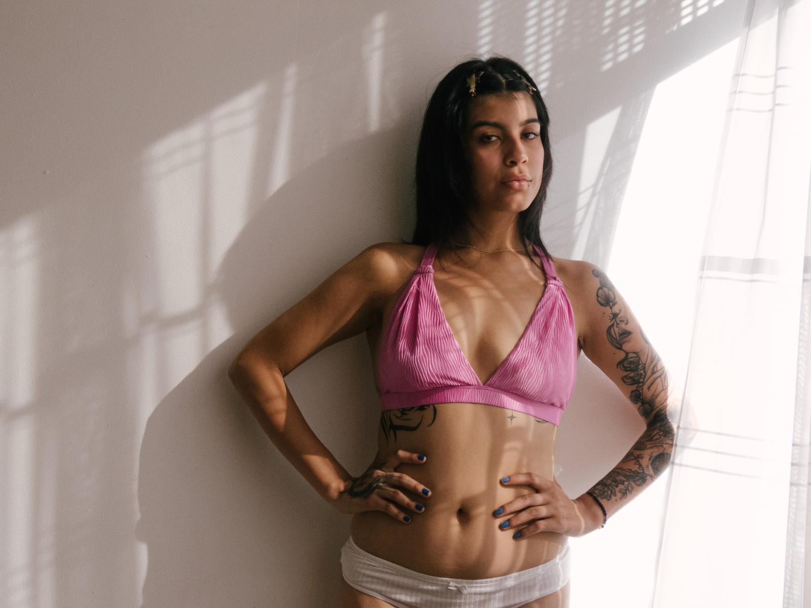 Fotografia de PsychesLoli em pé contra uma parede branca, com a luz do sol projetando sombras. Ela está vestindo um sutiã de renda rosa e calcinha branca, com as mãos nos quadris, e tem tatuagens no braço direito. Ajustado, sexy, confiante.