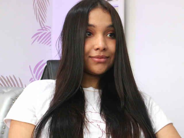 Photograph of BelleLane with long, straight black hair, wearing a white t-shirt, sitting against a white background with faint leaf patterns. She has a slight smile, dark eyes, and a relaxed pose. Her hair partially covers her chest.