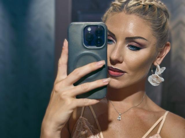 Close-up photo of FortyLeride, blonde with braided hair, holding a black smartphone, taking a selfie. She's wearing a beige lace top, red lipstick, and large white earrings. Her skin is shiny.