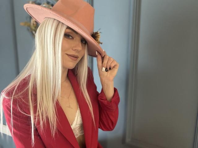 Fotografia di FortyLeride con lunghi capelli bianchi, che indossa un cappello rosa, una giacca rossa e una camicetta bianca in pizzo. Lei tiene il cappello con un sorriso malizioso, mostrando le unghie nere. Assolutamente attraente..
