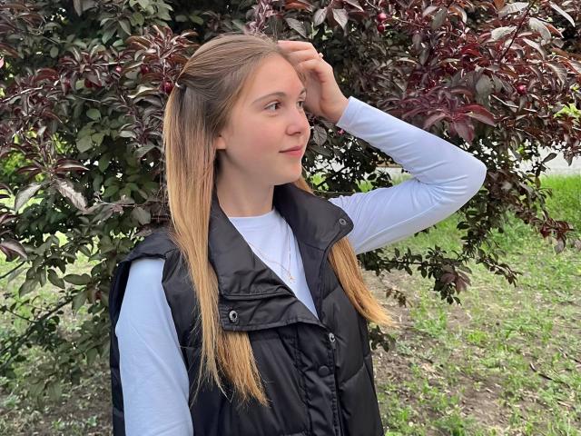 Deeann Milloy è in piedi di fronte a una vegetazione rigogliosa, verde e rossa, indossando un gilet nero sopra una camicia bianca. Ha lunghi capelli biondi, con una mano tra i capelli, e guarda leggermente di lato. Molto attraente..