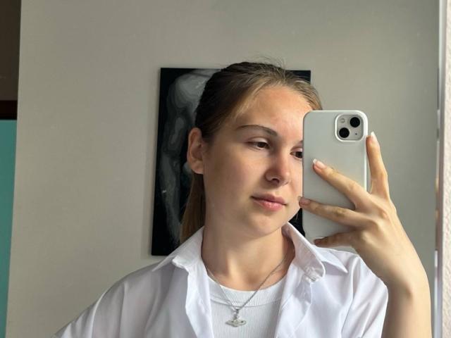 Deeann Milloy macht ein Selfie mit ihrem weißen iPhone, trägt ein weißes Hemd und eine weiße Halskette und hat helle Haut. braunes Haar im Pferdeschwanz und ein leicht verführerischer Ausdruck..