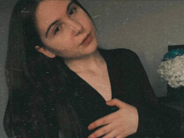 Fotografía de Deeann Milloy, de piel clara y cabello largo y oscuro, vestida con una blusa negra, con una mano en el pecho, expresión suave, de color oscuro. Fondo, ligera textura granulada, flor blanca en un jarrón en el lado derecho. Increíblemente atractivo/a..