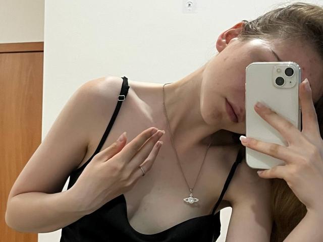 Deeann Milloy, sosteniendo un iPhone, se toma un selfie frente al espejo. Lleva un vestido negro con tirantes que deja ver parte del escote, y un collar de diamantes. Su mano cubre su pecho, mostrando tensión sexual. Iluminación brillante..