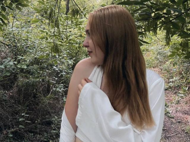 Deeann Milloy, une femme à la peau claire avec de longs cheveux bruns, se tient dans une forêt, vêtue d'un haut blanc à une épaule. Elle tourne la tête et affiche un sourire joueur et séduisant. Fond vert avec des feuilles..