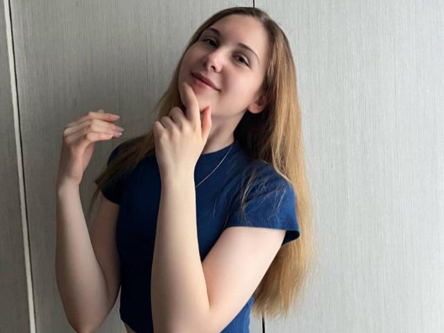 Deeann Milloy, com cabelos loiros longos e lisos, vestindo uma camiseta azul, em pé em frente a uma parede branca com textura, levemente... tocando o queixo com a mão direita, sorriso suave, pose casual, luz natural..