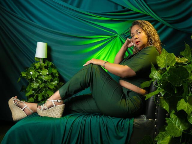 ManyHawkey, a curvy woman with light brown skin and curly hair, lounges on a black chair, wearing black pants and white wedge sandals, with green and black drapery. She's sexy, looking at the camera.