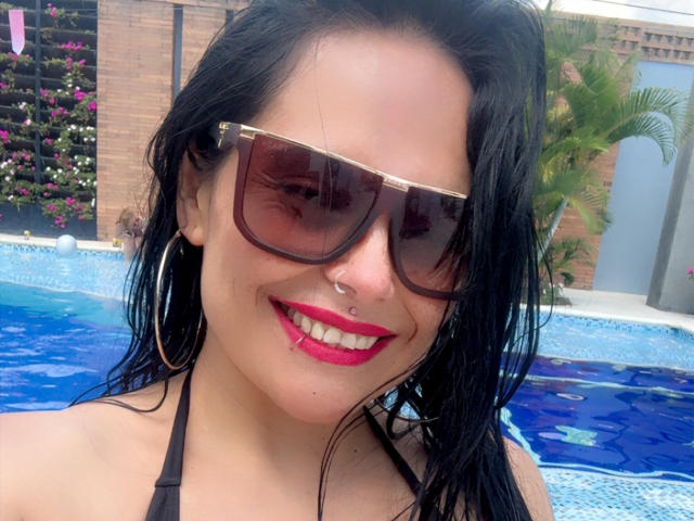 Close-up of CharlotteCrofort, wet black hair, wearing large brown sunglasses, red lipstick, black bikini, hoop earrings, nose ring, smiling at the camera, blue pool in background, brick wall. Fucking hot.