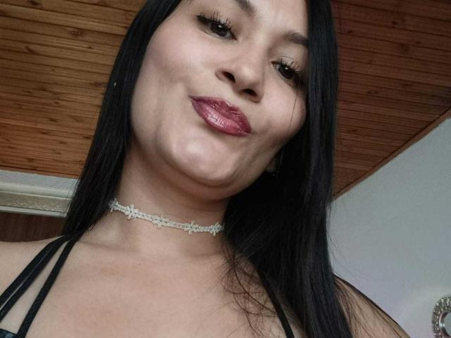 Fotografía de primer plano de KendallBrzz con cabello negro largo, labios rosados brillantes y un collar llamativo. Está sonriendo ligeramente, con un toque de tensión sexual, y lleva un top negro con tirantes..