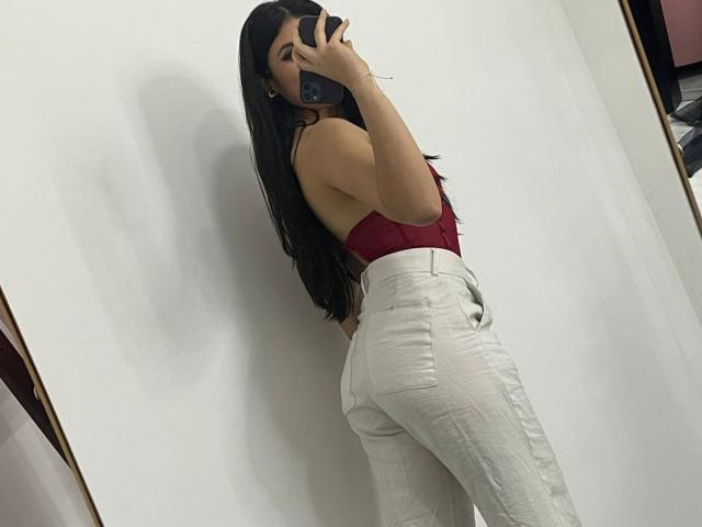 MiluSky maakt een selfie, waarbij ze over haar schouder kijkt. Ze heeft lang zwart haar, een rood topje zonder bandjes en witte broek met hoge taille, waardoor haar ronde billen benadrukt worden. Haar telefoon bedekt haar gezicht..
