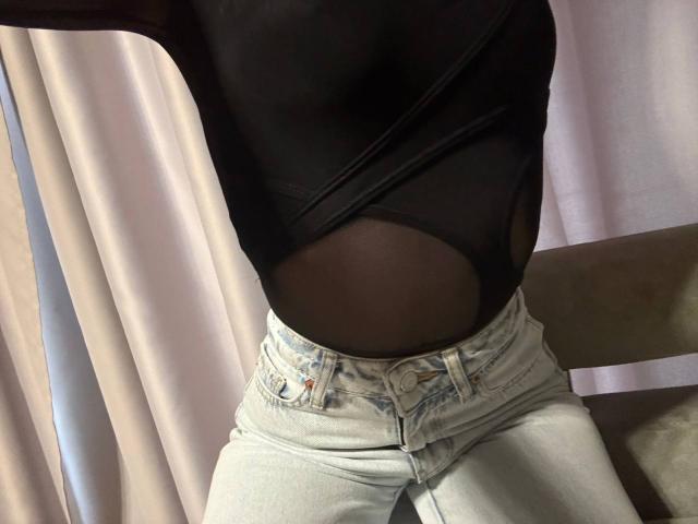 Esfil wears a black shirt with a visible belly button, light blue denim shorts. Sunlight through curtains highlights the waistline. Esfil's pose is suggestive, with a hint of sexual tension.