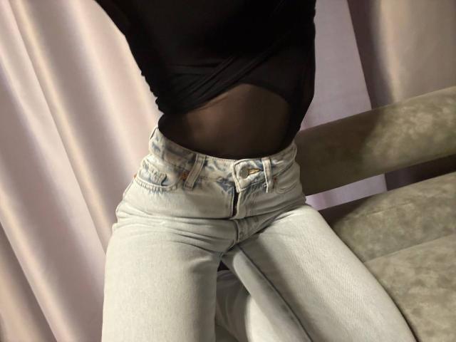 Primer plano de la parte inferior del cuerpo de Esfil, vistiendo unos vaqueros ajustados de color gris claro con costuras visibles y una camiseta negra que deja ver un cuerpo tonificado. abdomen. La mano de Esfil descansa sobre un sofá color beige, creando una pose provocativa..