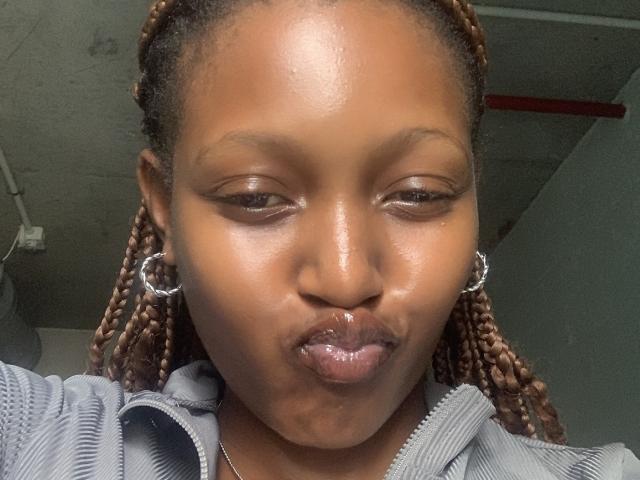 Foto em close-up de YumekoHinata, uma mulher negra com pele brilhante, cabelo trançado, usando brincos de argola prateados e camisa listrada cinza. Ela está fazendo beicinho com seus lábios cheios, criando uma expressão divertida e ligeiramente sensual..