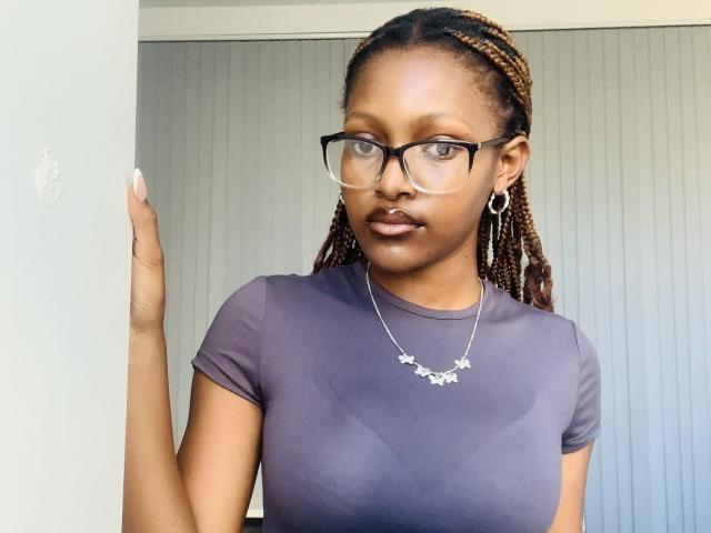 YumekoHinata stands in front of a vertical paneled wall, wearing a tight, dark purple t-shirt that outlines her breasts. She has braided hair, glasses, and a star necklace. Fucking sexy.