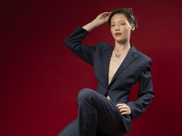 LissyLuxe, een vrouw met een lichte huid en kort zwart haar, draagt een donkerblauwe blazer, gedeeltelijk open om een onbedekte een borst en een ketting. Ze zit, met haar hand in haar haar, op een rode achtergrond..