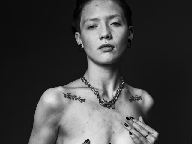 Fotografía en blanco y negro de LissyLuxe, sin sujetador y con tatuajes en los hombros, vistiendo un collar de cadena, con una mano cubriendo el lado izquierdo. pecho, fondo oscuro. Alto contraste, mirada intensa, esmalte de uñas negro, sensualidad..