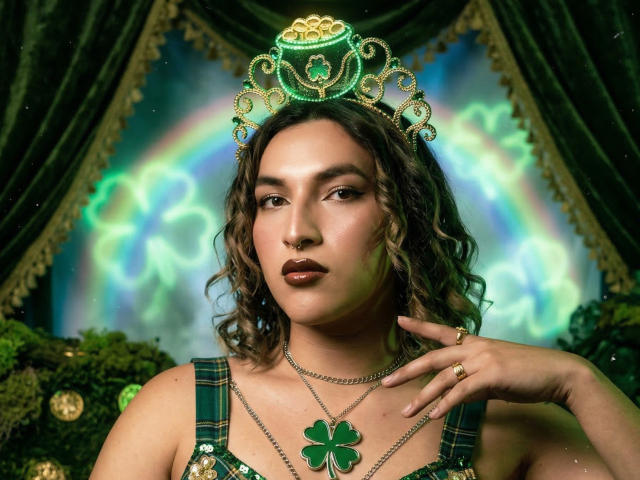 TommyTS, con i capelli castano ondulati, indossa una collana con un quadrifoglio verde e una corona elaborata. Luci verdi brillanti e tende scure sullo sfondo. La mano di TommyTS tocca il collo, in modo seducente. Aura luminosa.