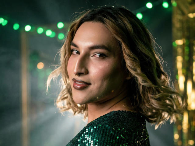 Close-up photo of TommyTS, a sexy woman with wavy blonde hair, wearing a black sequined top, illuminated by green and yellow bokeh lights. Her lips are slightly parted, exuding confident allure.