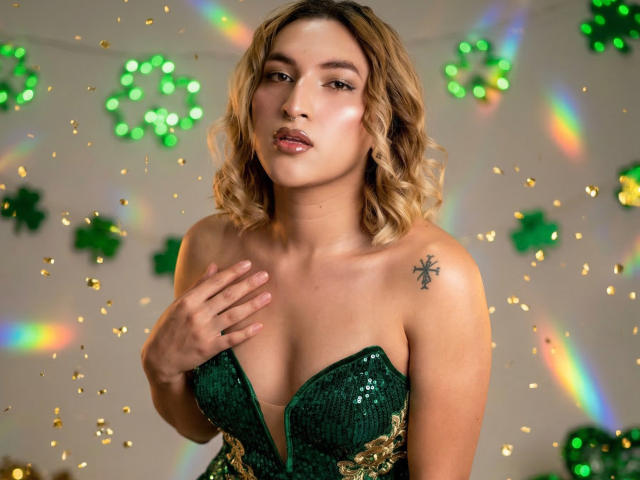 TommyTS, com cabelos loiros ondulados, veste um vestido verde sem alças, coberto de lantejoulas, com a mão no peito, um leve sorriso e uma tatuagem de estrela preta. no ombro esquerdo, fundo colorido com efeito bokeh, formas verdes brilhantes..