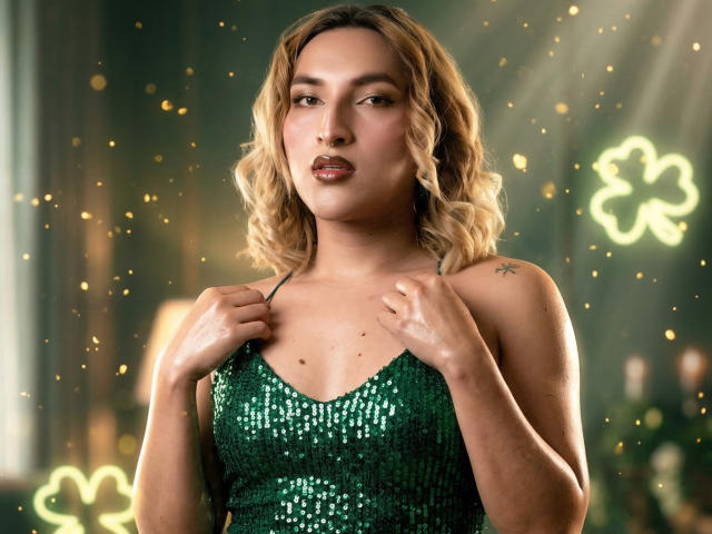 TommyTS se tient fièrement dans une robe verte ornée de paillettes, les mains agrippant les bretelles, avec des fleurs néon lumineuses dans les contexte. La lumière se reflète sur les paillettes, mettant en valeur ses courbes et son expression sensuelle..