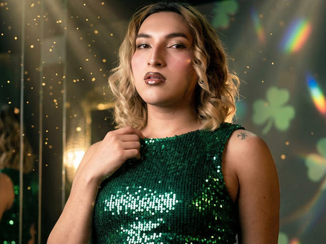 TommyTS, con cabello rubio ondulado, vestida con un vestido verde con lentejuelas, sosteniendo el escote. Fondo brillante con reflejos de arcoíris. Labios brillantes, mirada ligeramente seductora, tatuaje en el hombro derecho. Fuente de luz brillante.