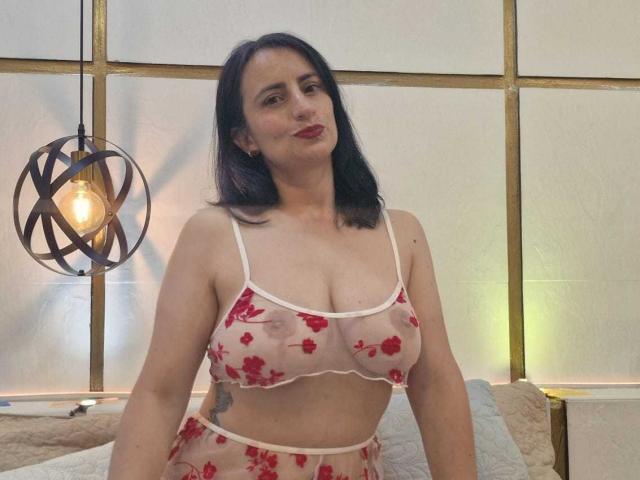 Brenda Cantorel, met zwart haar dat tot op haar schouders reikt, draagt een doorschijnende witte beha met een rood bloemenpatroon, waardoor haar borsten van gemiddelde grootte en een tatoeage op de linkerarm. Warm licht van de geometrische hanglamp, sensuele pose.