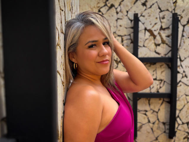 Photograph of CamilaNoer with medium brown skin, shoulder-length blonde hair, wearing a pink strapless dress, leaning against a textured stone wall, right hand behind her head, smiling slightly.