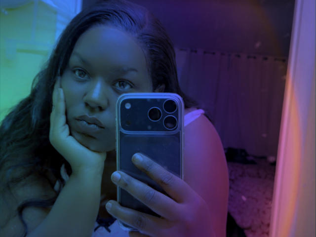 EbonicQueen takes a mirror selfie with a blue and green-lit phone. She rests her hand on her cheek, gazing intensely into the camera. Her dark hair cascades over her shoulders.