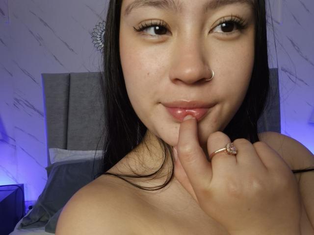 Primer plano de ScarllettDream, con piel clara, cabello negro y un piercing en la nariz, mordiéndose el dedo de forma seductora. Ella tiene labios carnosos, ligeramente entreabiertos, y lleva un anillo de oro en el dedo. Fondo con iluminación azul..