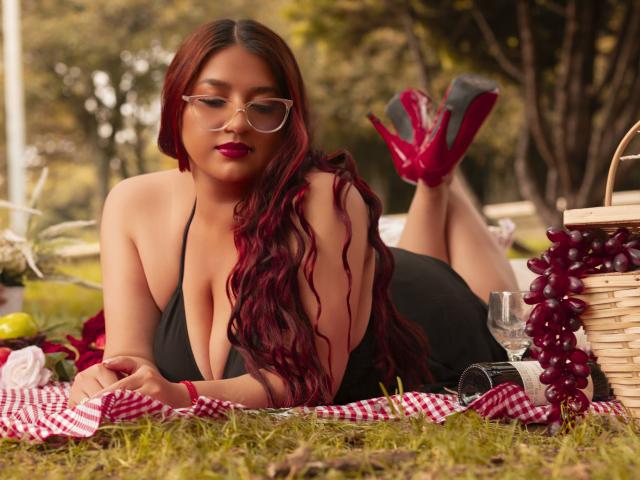 Tiana è sdraiata su una coperta da picnic, con le gambe sollevate, indossando un top nero a tracolla, tacchi rossi e occhiali. Ha lunghi capelli rossi, rossetto rosso e un'espressione sexy e seducente..