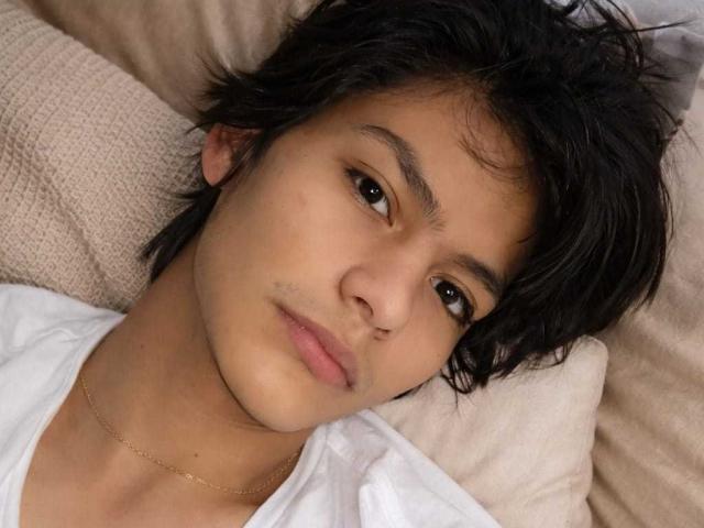 Primer plano de AxelPrince con el pelo oscuro y despeinado, acostado sobre un cojín beige, vistiendo una camisa blanca y una cadena de oro. Mirada suave y sensual, mejillas ligeramente sonrojadas, expresión relajada. Ambiente relajado e íntimo.