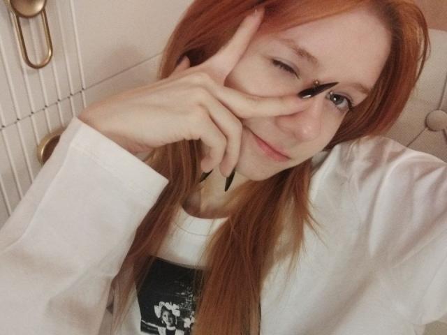 Gros plan de JamieLathrum, une jeune femme aux longs cheveux roux, qui cligne de l'œil et fait un signe de la paix avec sa main droite, portant une chemise blanche, un sourire suggestif, sexy, une expression coquine..