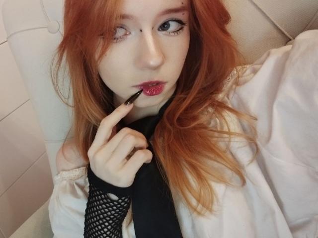 Jamie Lathrum, com pele clara e cabelo ruivo, mordendo um aplicador de batom preto, com olhos arregalados, vestindo uma camisa branca e preta. manga de meia-calça, aparência sedutora, colar no pescoço, olhar intenso, sensualidade..