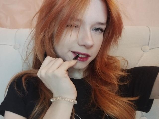 Foto em close-up de JamieLathrum, uma ruiva de pele clara e cabelos longos, mordendo o dedo de forma sedutora, vestindo preto uma camisa e um bracelete, com uma expressão provocante e sensual, lambendo os lábios..