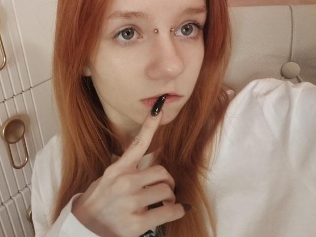 Nahaufnahme von JamieLathrum, einer blassen Frau mit roten Haaren, die sich die Nase piercen lässt und dabei einen schwarzen Stift zwischen die Zähne nimmt. Zeigefinger, trägt ein weißes Hemd, vermittelt eine provokante, sexuelle Ausstrahlung..