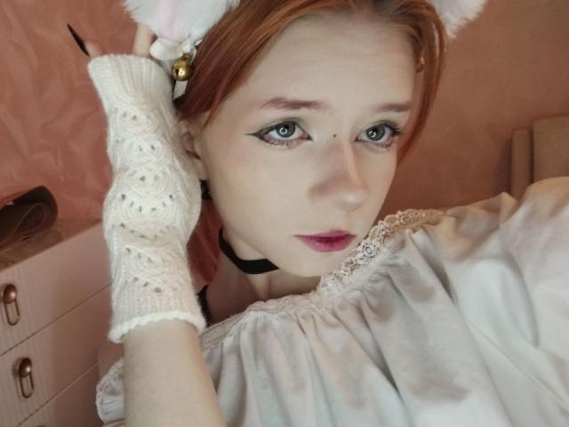 Fotografia ravvicinata di JamieLathrum, con la pelle chiara e i capelli rossi, che indossa guanti di pizzo bianco, orecchie da gatto e un collare. Lei guarda in avanti con gli occhi azzurri, le labbra leggermente dischiuse, rivelando una sottile tensione sessuale..