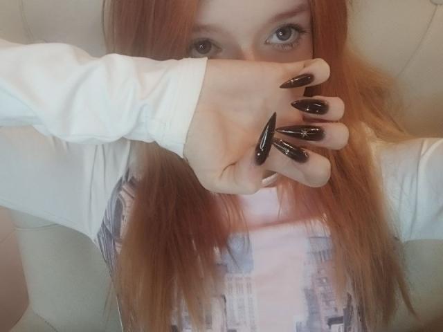 Photo en gros plan de JamieLathrum, à la peau claire et aux longs cheveux roux, qui se couvre le visage avec sa main et dont les ongles noirs et brillants sont visibles. portant un haut blanc, motif urbain, regard intense, légère tension sexuelle, ambiance séduisante..