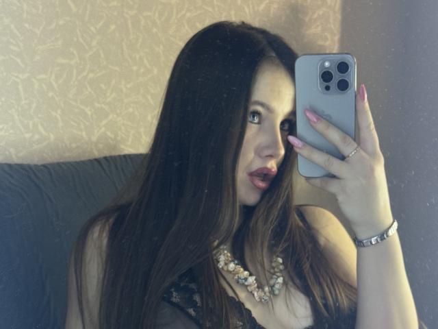 BarnyHot, avec ses longs cheveux bruns et son rouge à lèvres rouge, prend un selfie devant un miroir avec un iPhone. Elle porte un haut noir, un collier de perles et un bracelet en argent, et elle a l'air séduisante. Incroyablement sexy.