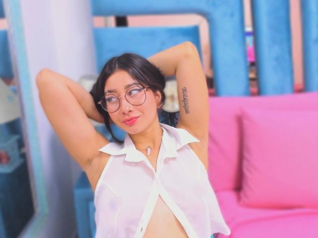 Photograph of Genesiss, a curvy woman with glasses, black hair, and a white sleeveless shirt, arms raised, showing a tattoo on her right arm. Bright blue and pink background. Fucking sexy.
