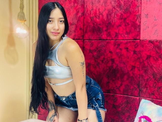 Emma Taylor se penche contre un mur rouge texturé, vêtue d'un crop top argenté et de shorts en jean. Longs cheveux noirs, tatouage sur son bras droit, sourire séduisant. Éclairage vif, pose aguicheuse..