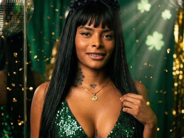 Fotografia di EmmaEbony, una donna di carnagione scura con lunghi capelli neri, che indossa un abito verde con paillettes, con un piccolo tatuaggio a forma di cuore su petto, collana d'oro, abito leggermente aperto, luci e scintille sullo sfondo.