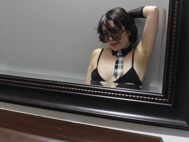 Photograph of ElodiePerrine with short black hair, glasses, and a black bra, wearing a plaid tie, leaning against a mirror with her arm raised, looking seductive. Reflective, dark-framed mirror.